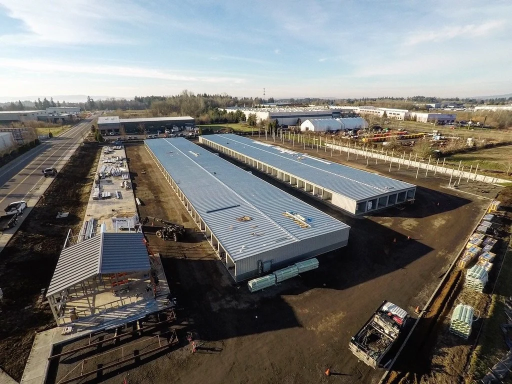 An aerial view of a construction site with multiple industrial buildings with metal roofs, surrounded by parking lots, cars, and trees in the background.