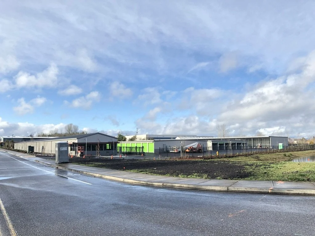 A construction site on a wet road shows a large building with a gray exterior, some green panels, construction cones, a portable toilet, and parked vehicles. The sky is partly cloudy.