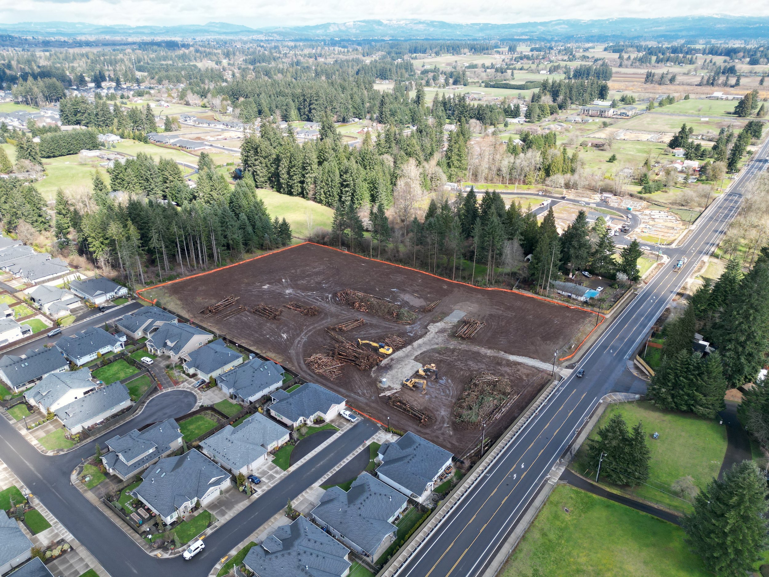 Aerial view of a cleared construction site bordered by a street and residential neighborhood with additional greenery and forests in the background.