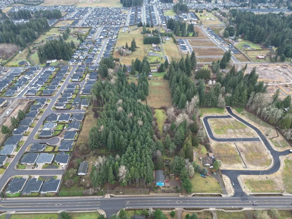 Aerial view of residential neighborhood with rows of houses on the left and a large wooded area in the center, surrounded by roads and some open plots of land.