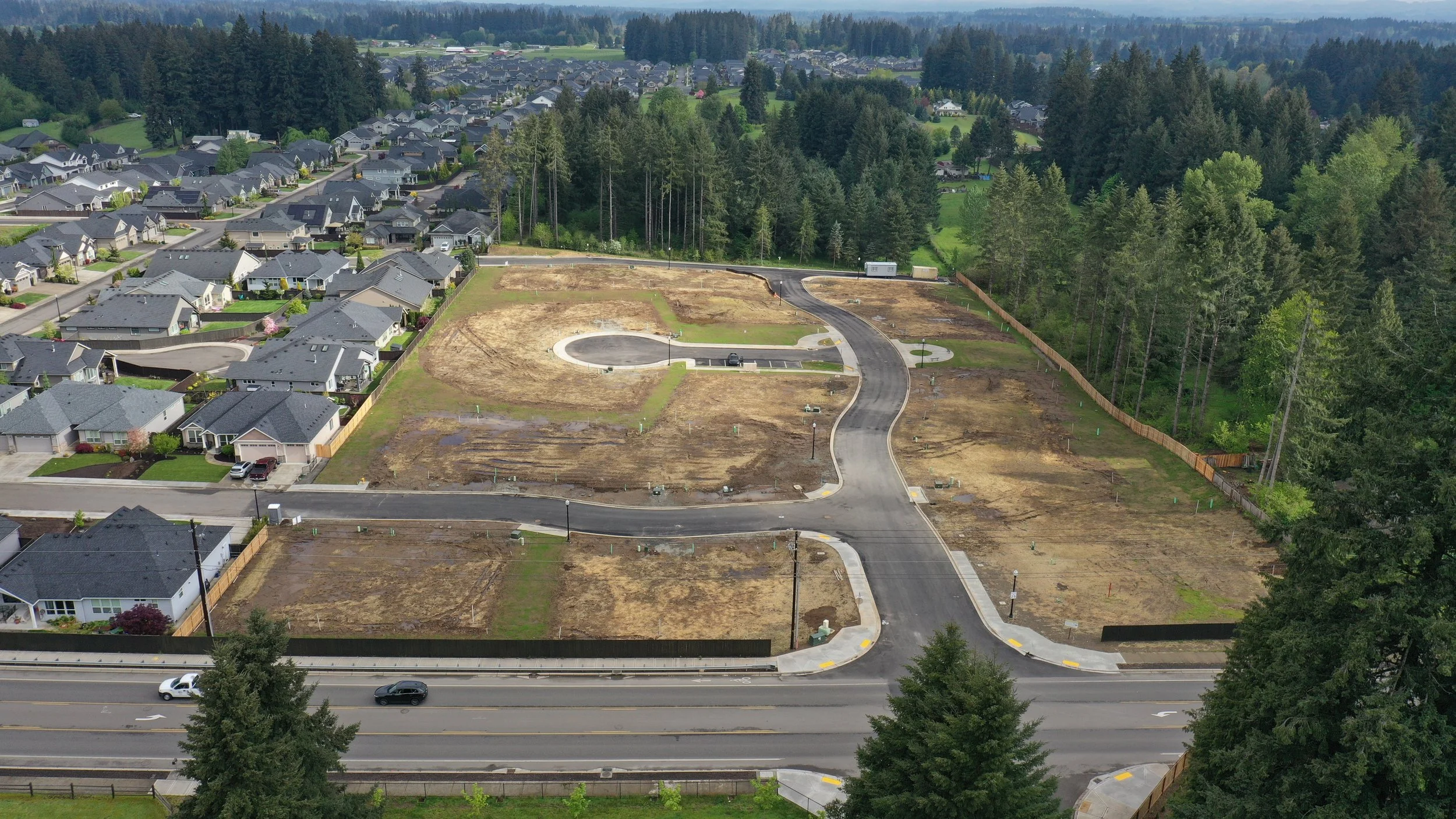Aerial view of a residential neighborhood with houses on the left and a developing area with cleared land, new roads, and some construction on the right, bordered by trees.