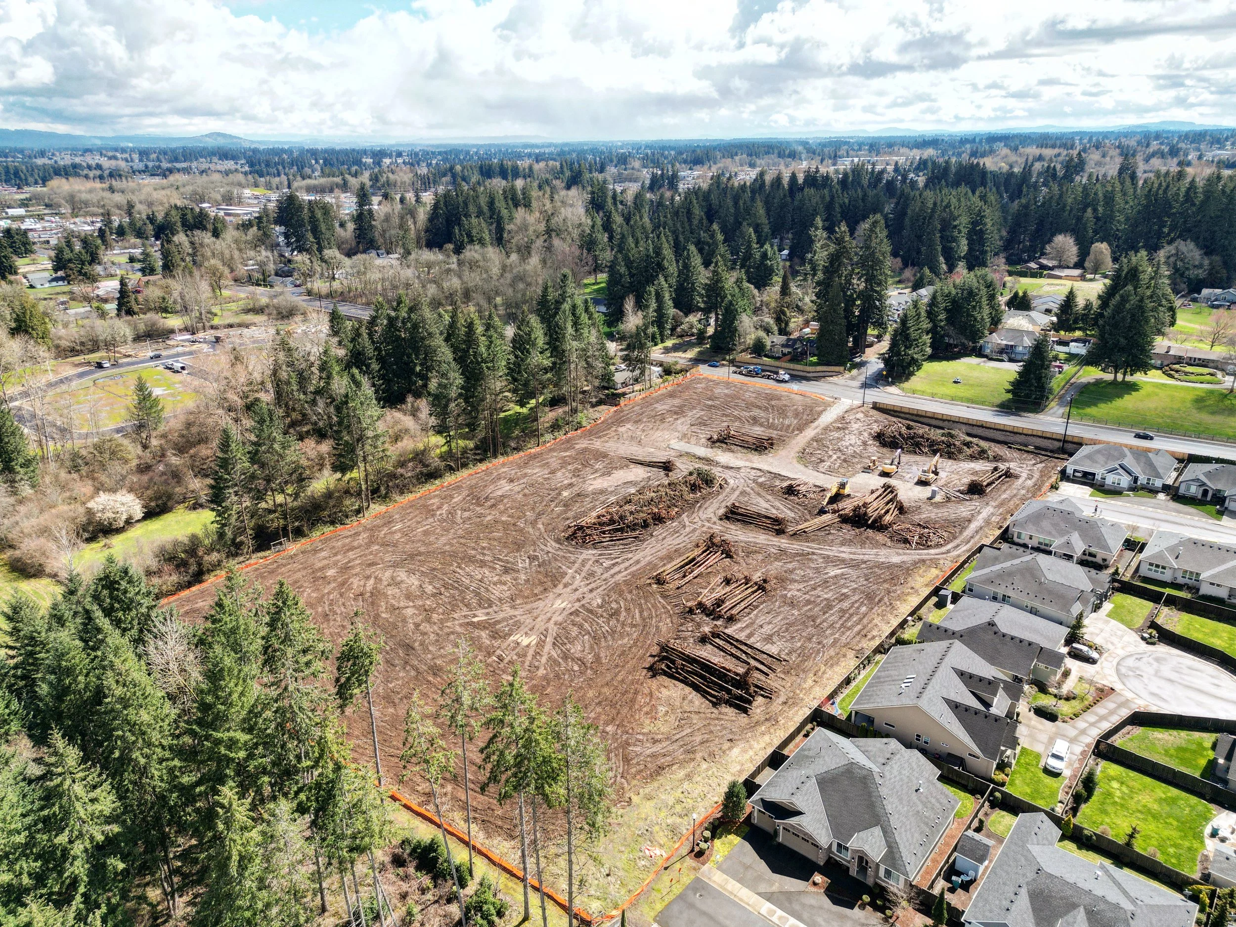 Aerial view of a cleared construction site with trees and residential houses nearby, under a cloudy sky.