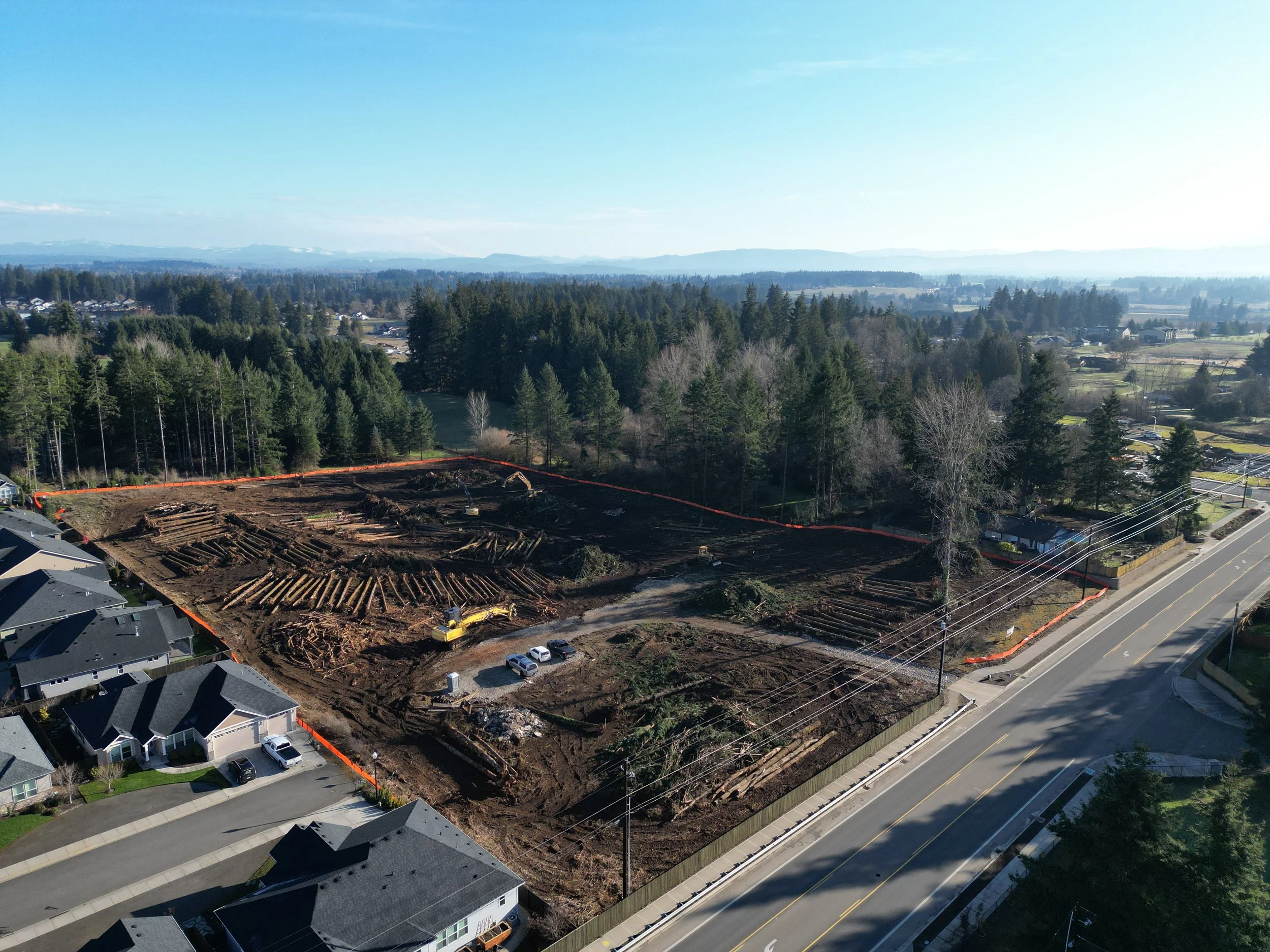 Construction site with cleared land, logs, and machinery, surrounded by residential houses and trees, in a suburban area with a road running alongside.