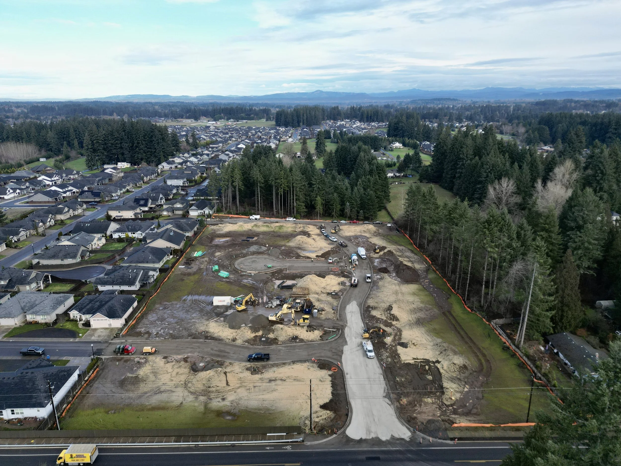 Aerial view of a construction site with machinery and vehicles, surrounded by residential houses and a forested area, under a partly cloudy sky.