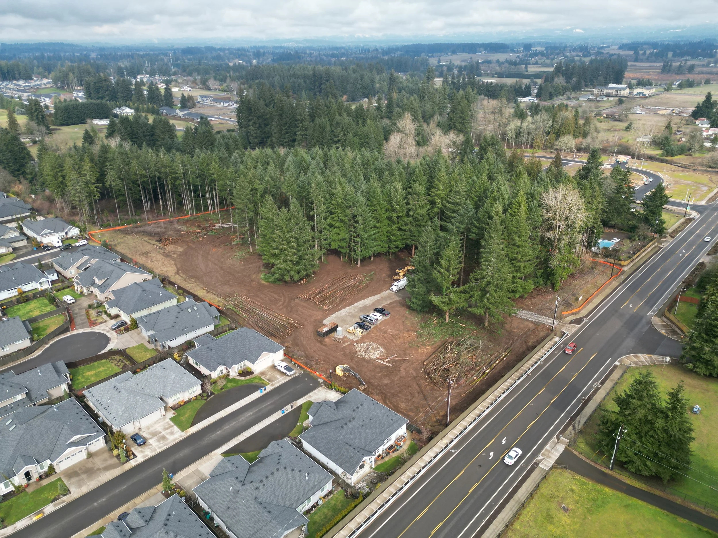 Aerial view of a residential neighborhood and a wooded area under construction near a highway, with trees and partly cloudy sky in the background.