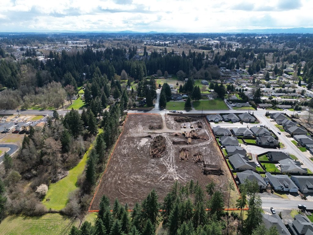 Aerial view of a construction site with cleared land, wooden logs, and equipment surrounded by residential neighborhoods and a forested area.
