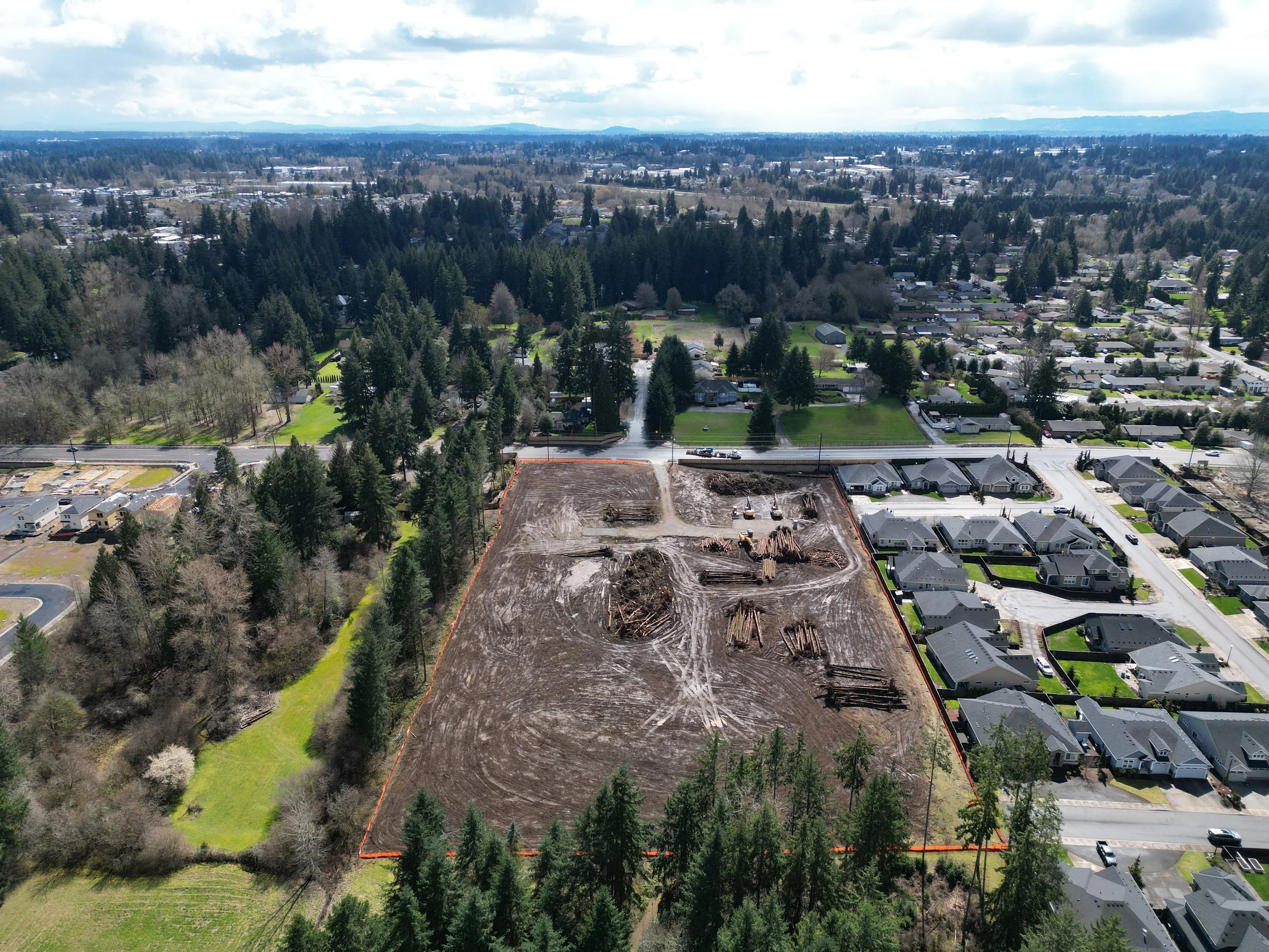 Aerial view of a cleared construction site bordered by trees and nearby residential houses on a cloudy day.