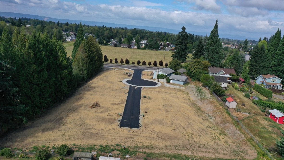 An aerial view of a newly paved driveway leading to a cul-de-sac in a suburban neighborhood, with houses, trees, and open fields surrounding the area.
