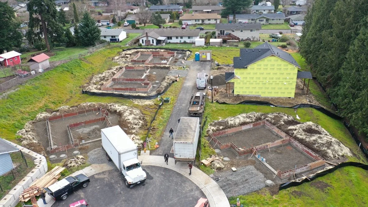 Construction site with several foundations being prepared for new buildings, surrounded by grassy areas and neighboring houses.