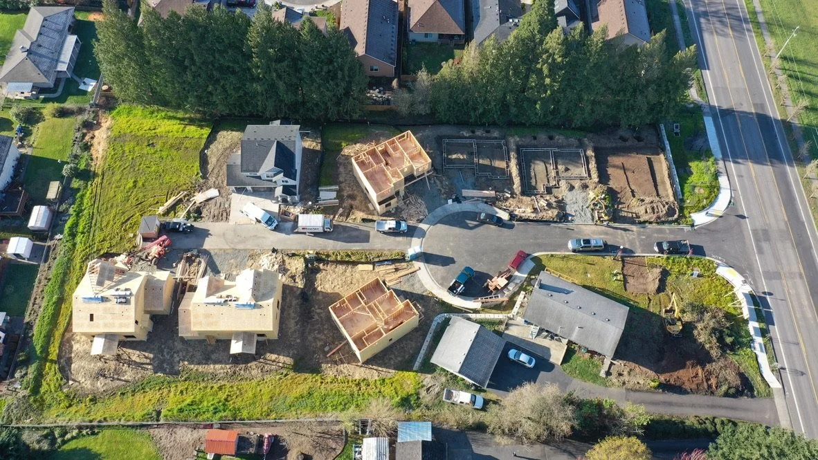 Aerial view of a residential construction site with partially built houses, construction vehicles, and surrounding trees and houses.