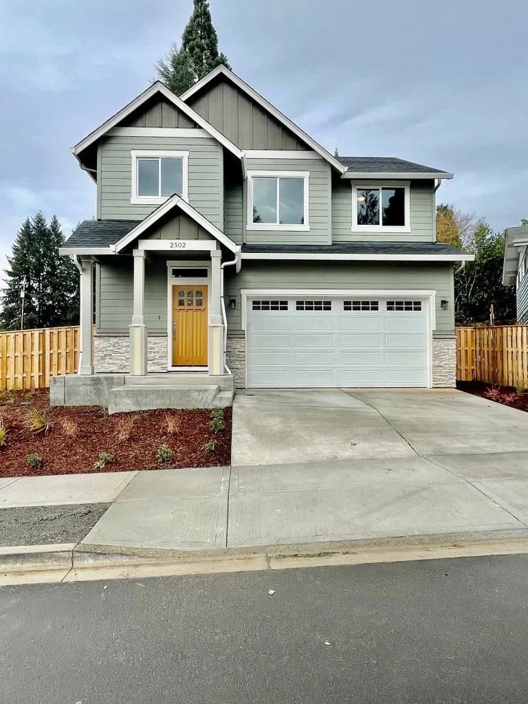 A modern two-story house with gray siding, a yellow front door, and a two-car garage, surrounded by a small landscaped yard and a wooden fence.