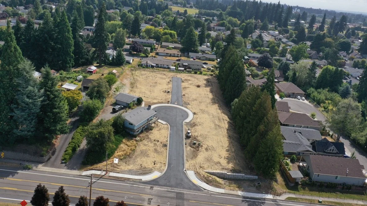 An aerial view of a residential neighborhood with a newly paved street leading to a cul-de-sac, surrounded by houses, trees, and open land.
