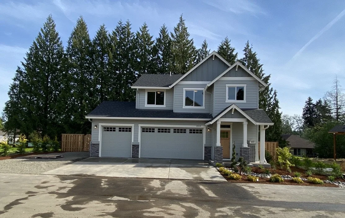 A two-story gray house with three car garage, front porch, and landscaped yard, surrounded by tall green trees.