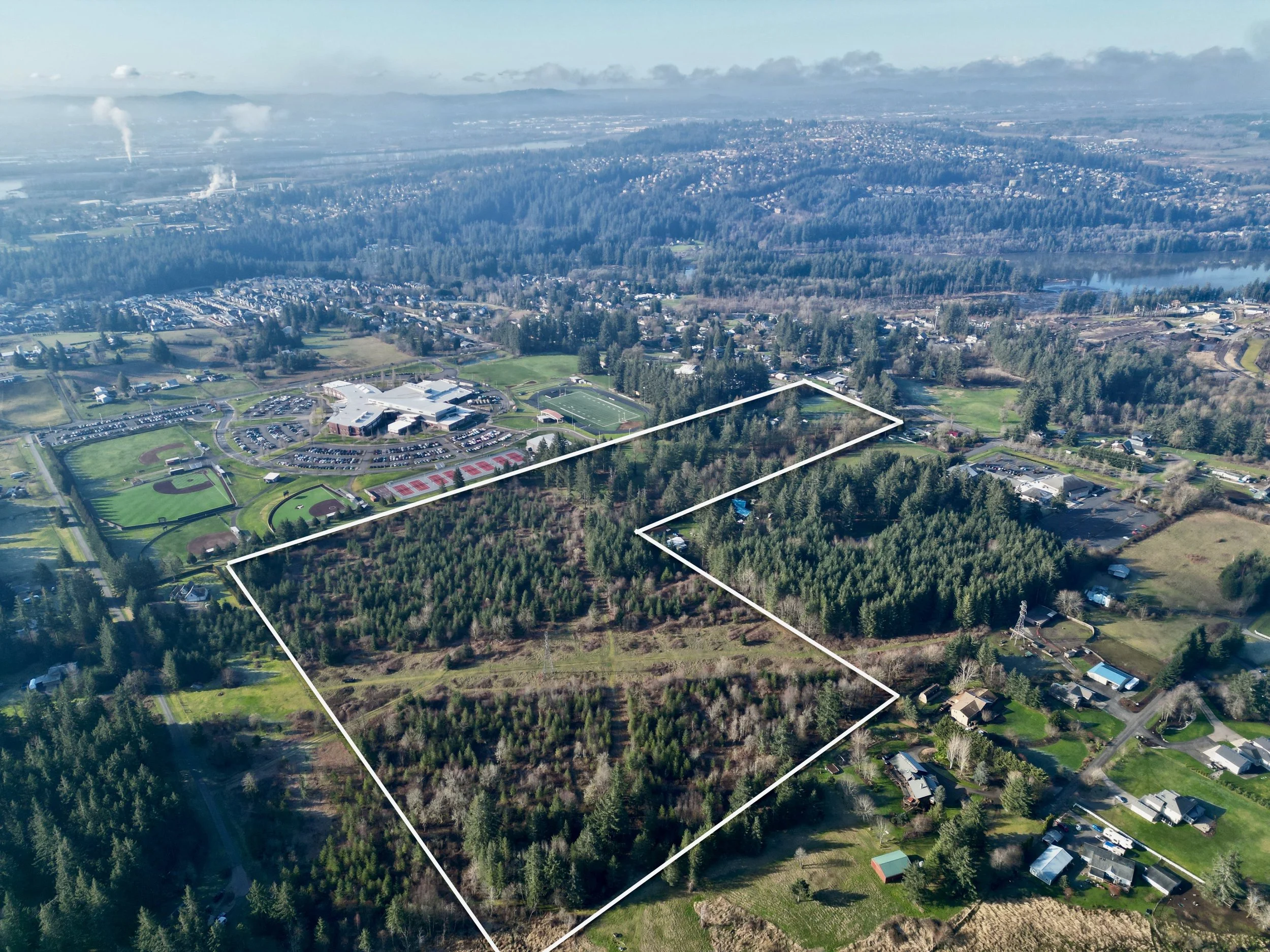 Aerial view of a large wooded plot of land outlined in white, surrounded by residential houses and a school with sports fields, with a body of water and forested hills in the background.