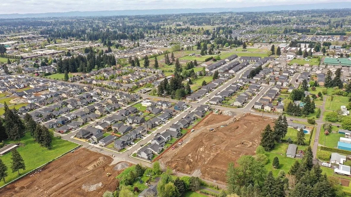 Aerial view of a suburban neighborhood with houses, streets, and green yards, including some areas under construction with dirt and equipment.