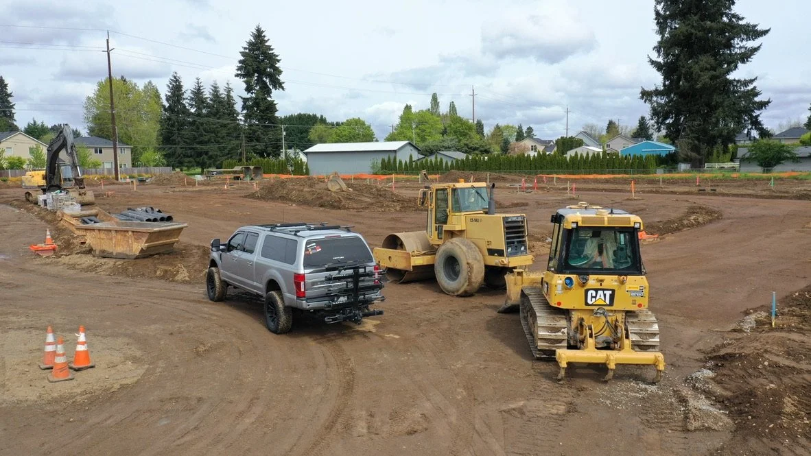 Construction site with heavy machinery including a bulldozer and a roller, dirt tracks, and orange safety cones, with residential houses and trees in the background.