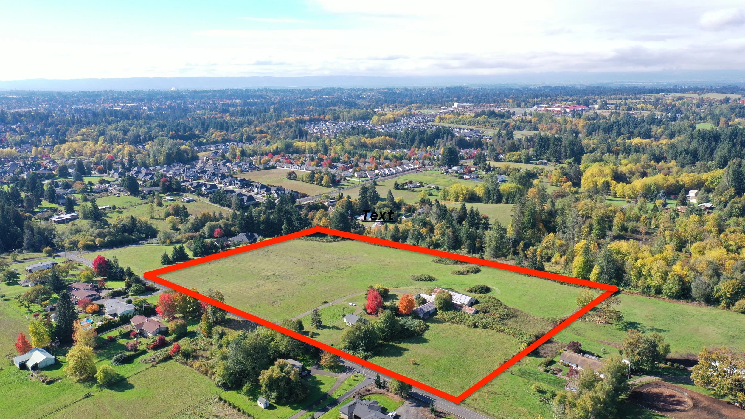 Aerial view of a large grassy lot outlined in red, with a few small buildings and trees, surrounded by residential homes and dense forest, with a city in the distance.