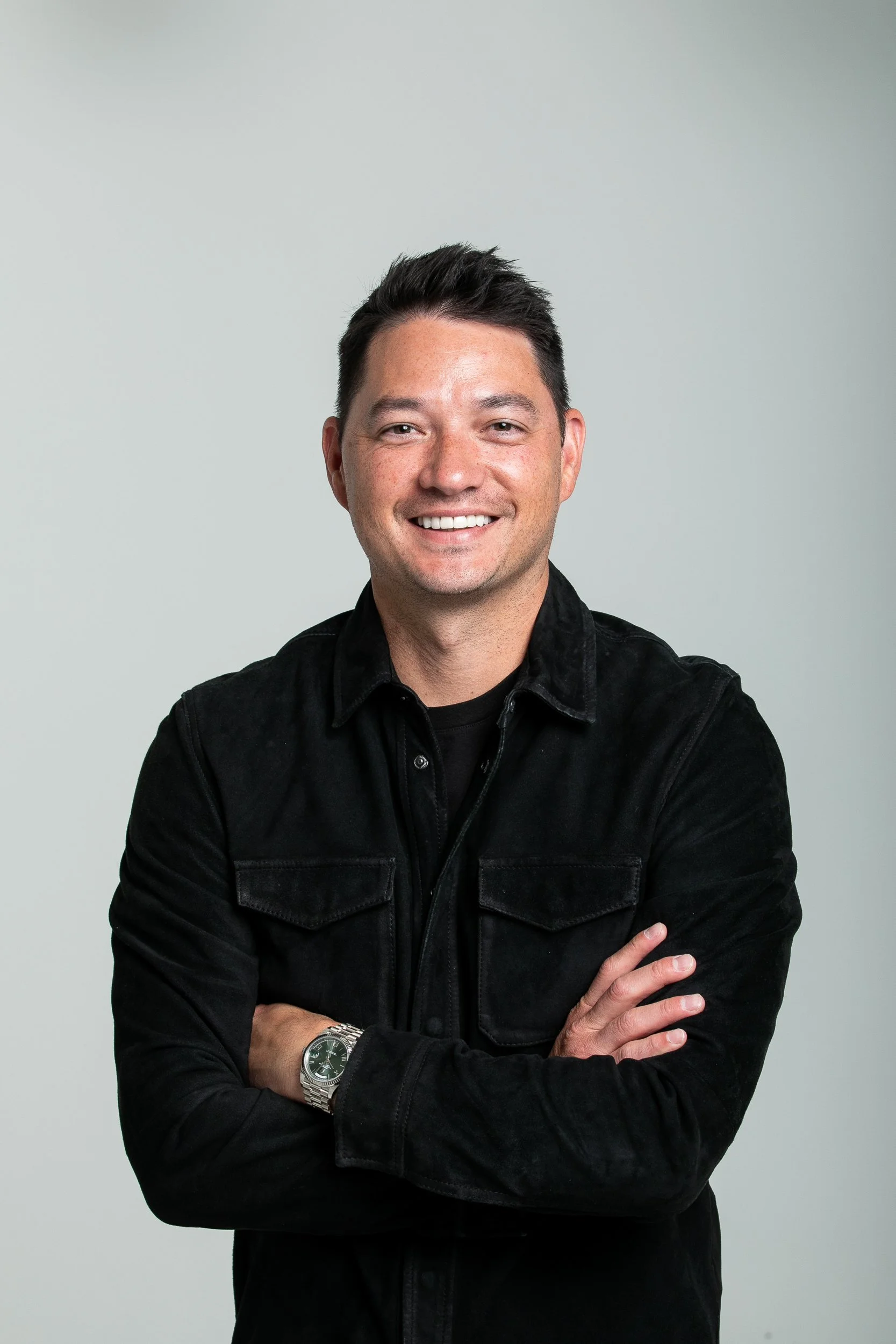 A smiling man with dark hair, wearing a black jacket and watch, crossing his arms against a light background.