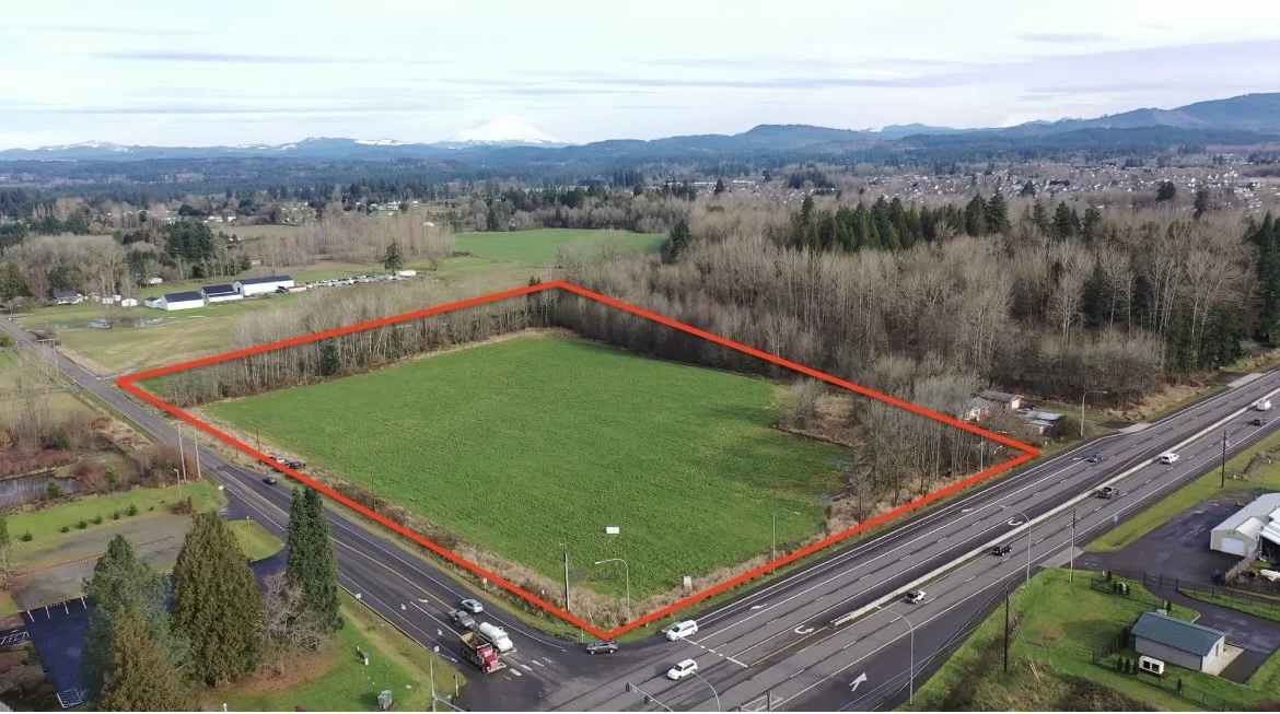 A rectangular piece of land outlined in red, situated at the intersection of two roads, with a green field inside and surrounded by trees, buildings, and distant mountains in the background.