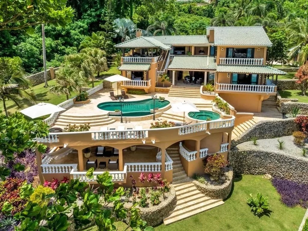 A luxurious multi-level house with multiple balconies and a swimming pool in the backyard, surrounded by lush greenery and tropical plants.