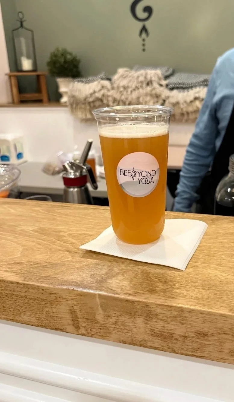 A plastic cup of light amber beer with a sticker that says 'BeeYOND Yoga' and a logo, sitting on a white napkin on a wooden bar or counter.