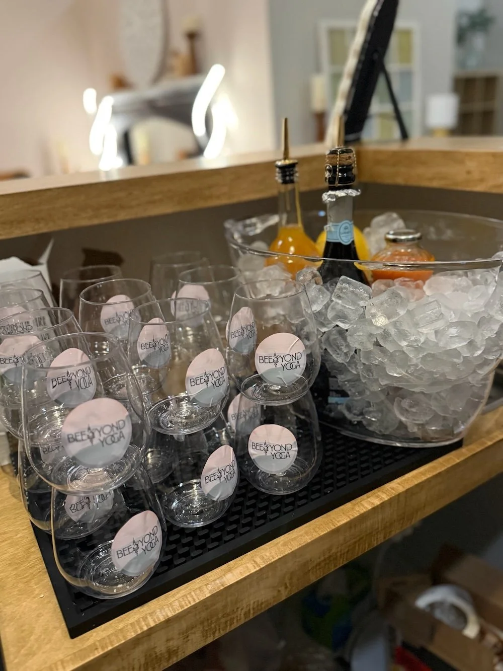 A wooden tray with a black rubber mat holds a large ice bucket filled with ice, bottles of champagne or sparkling wine, and some small glasses labeled 'BEEYOND YOGA'.
