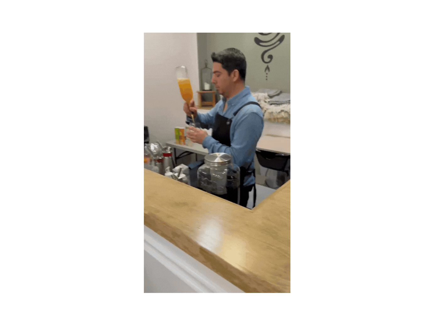 A male bartender in a blue shirt and black apron preparing a cocktail with an orange-colored drink in a tall flute glass at a bar counter, with various bar tools and accessories around him.