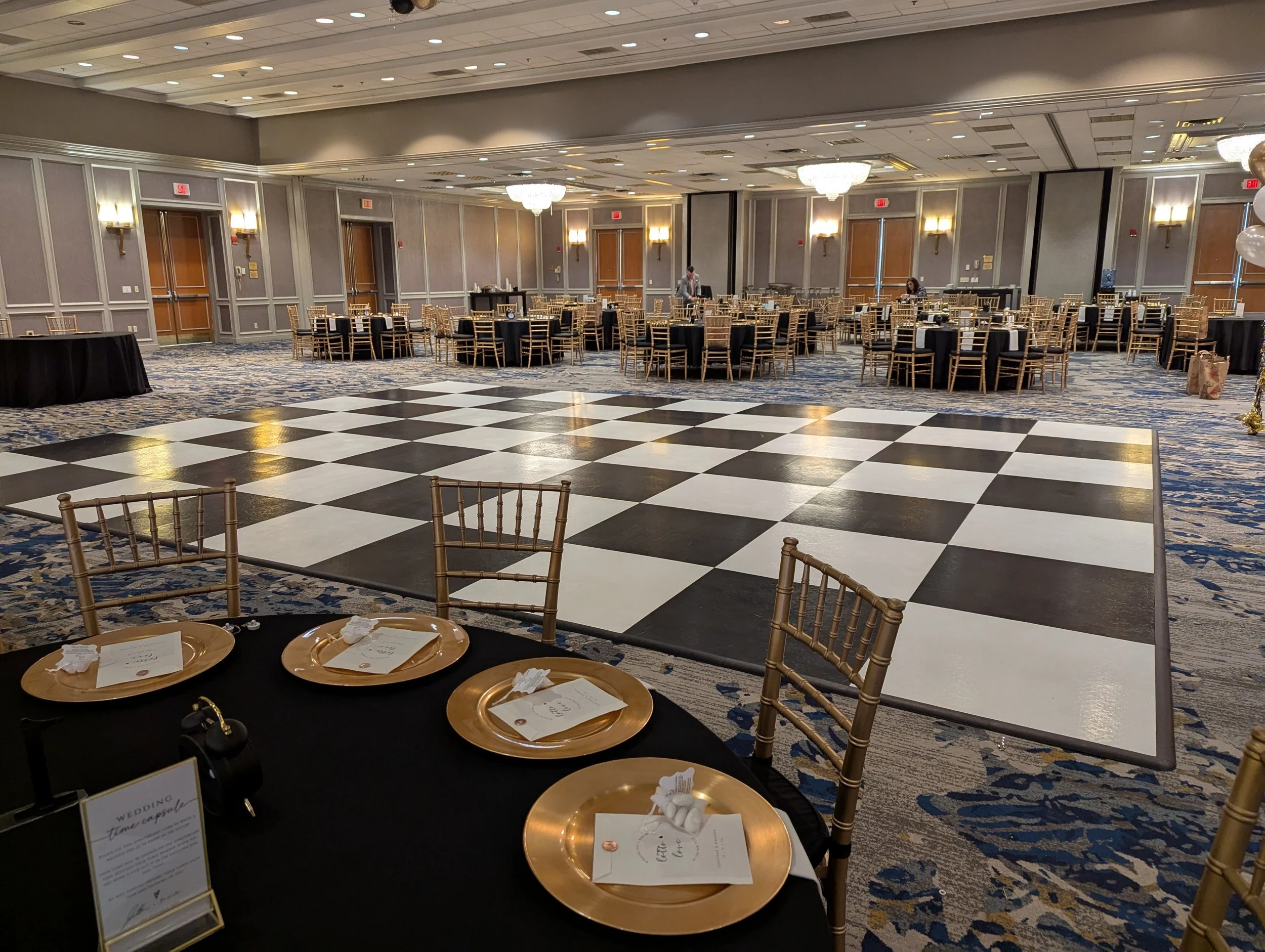 Empty banquet hall with a large checkered dance floor in the center, tables with black tablecloths and gold chairs, and a few people setting up or preparing.