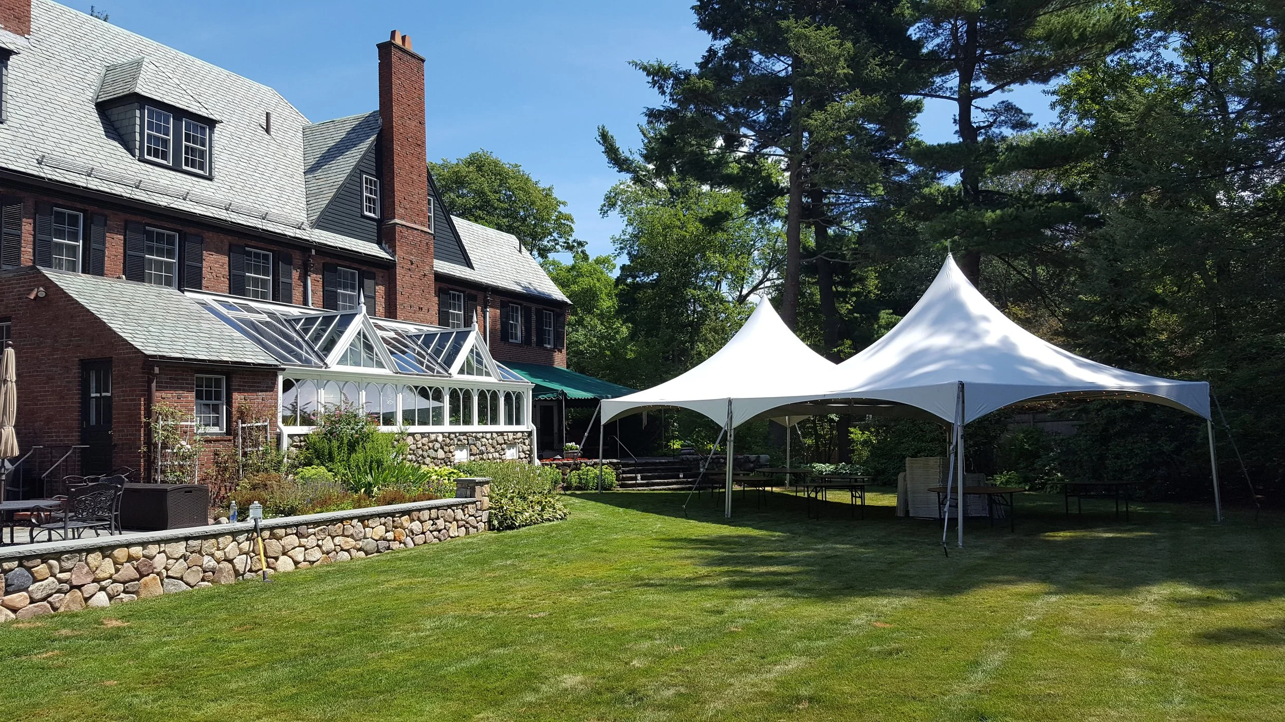 Large house with brick walls and a glass greenhouse attached to it, set in a landscaped garden with a grassy lawn and tall trees. Two white tents are set up on the lawn, likely for an outdoor event.