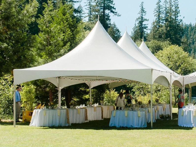 Outdoor event space with white tents, tables, and chairs set up on grass surrounded by trees and greenery.