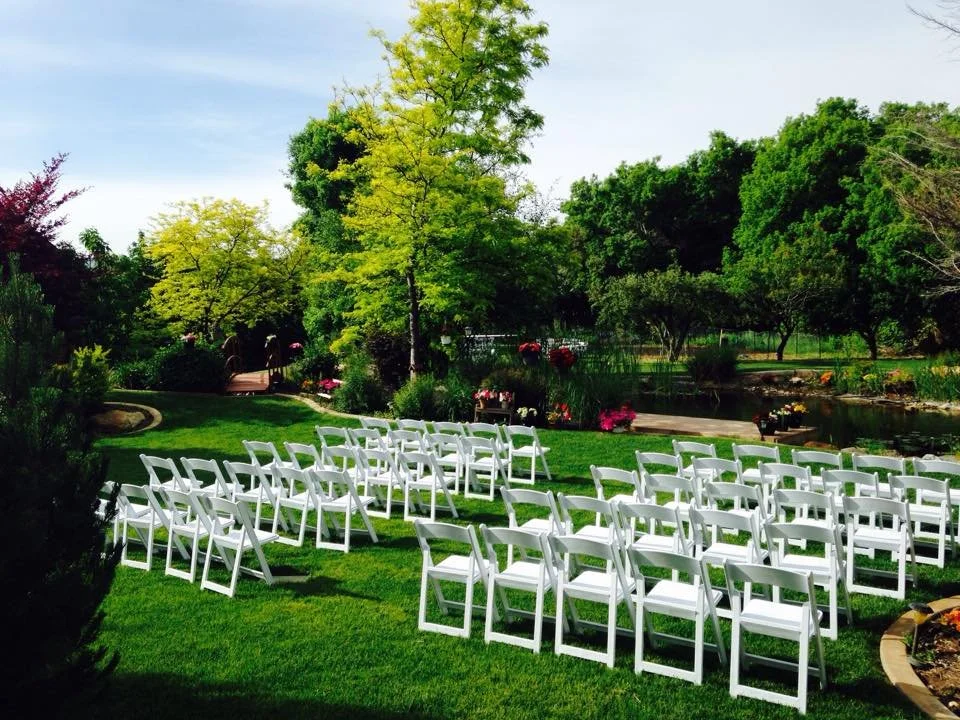 White folding chairs are arranged outdoors on a lush green lawn in front of a pond. The background features various trees and colorful flowers, suggesting a garden setting for a wedding or outdoor event.