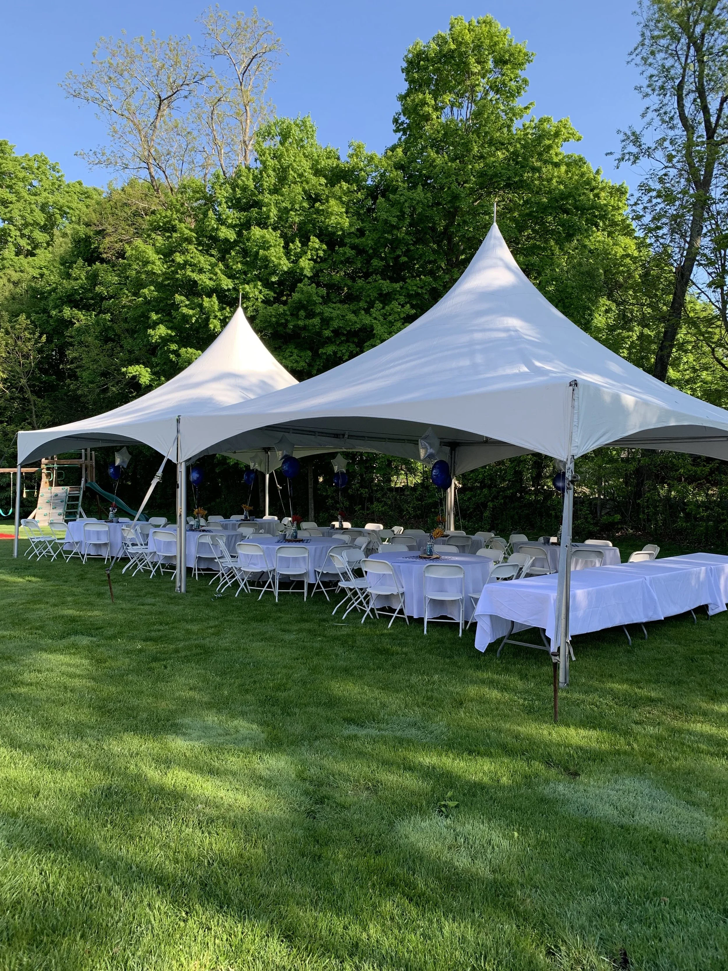 White event tent set up on a grassy area with round and rectangular tables covered with white tablecloths, surrounded by white chairs, with blue balloons hanging inside, and a playground in the background, surrounded by green trees against a clear bl