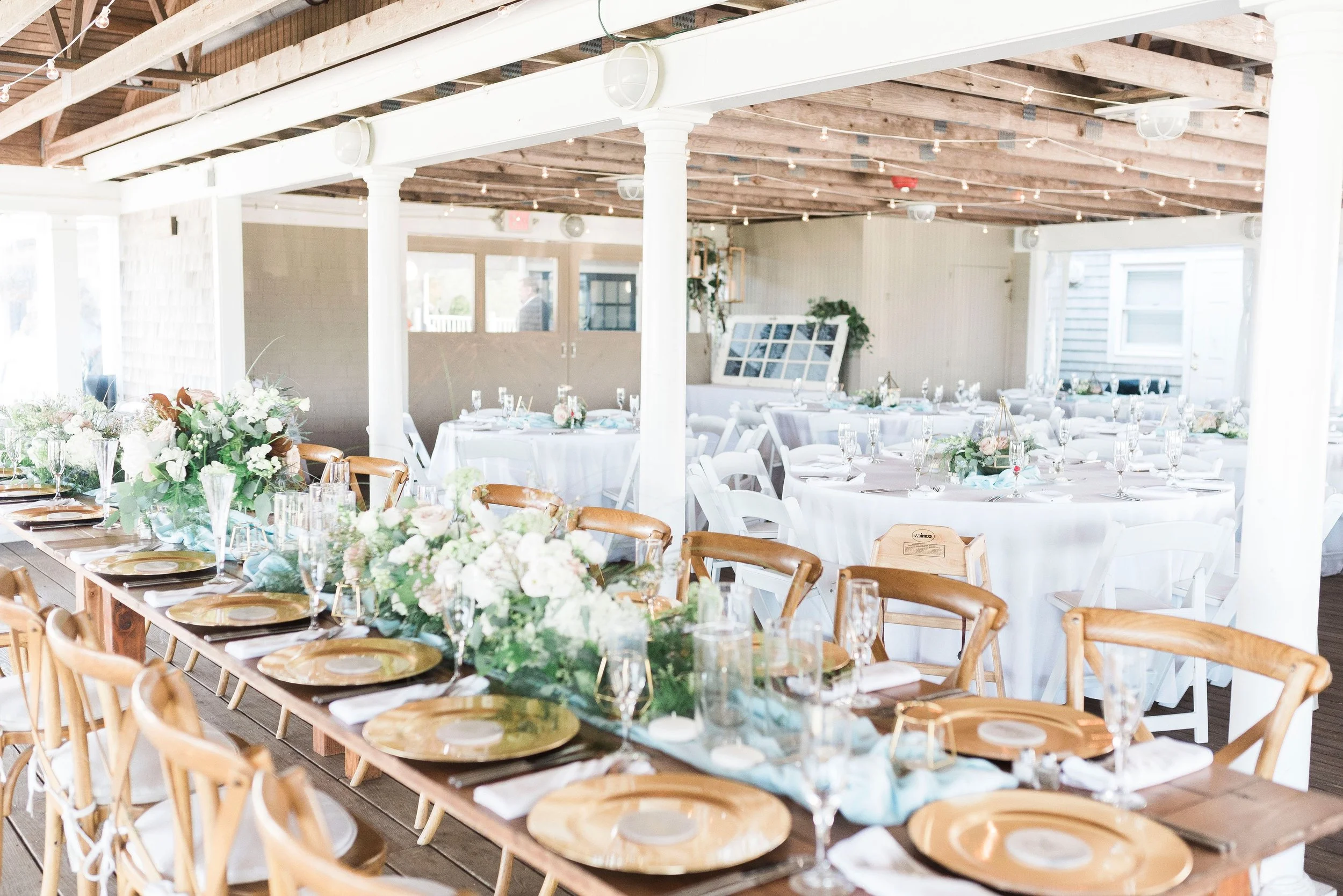 Wedding reception setup with long wooden table decorated with white and light pink flowers, gold plates, white napkins, and glassware, surrounded by white and wooden chairs inside a rustic venue with exposed wooden beams.