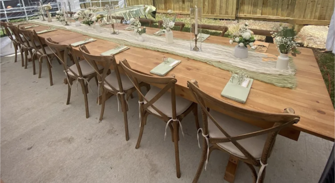 Long wooden table set for a gathering with folded napkins, flowers, candles, and decorative items, surrounded by wooden chairs with tied ribbons, in an outdoor space.