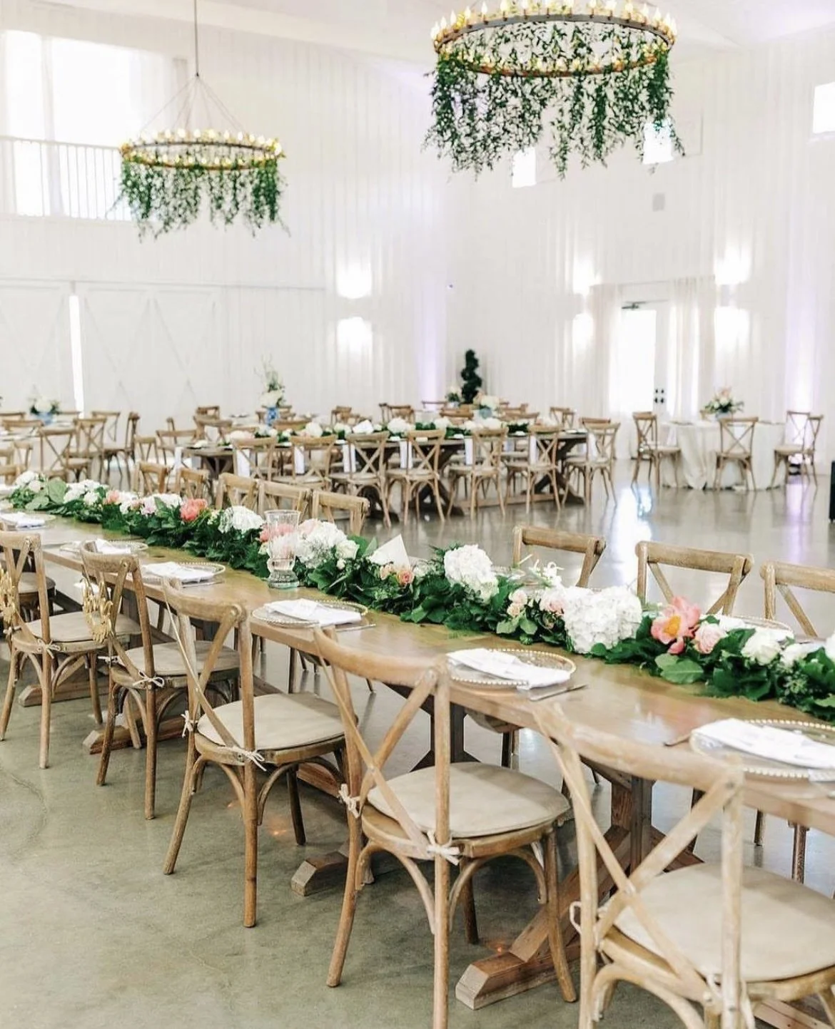 Elegant wedding reception venue with long wooden tables decorated with white and pink floral centerpieces, surrounded by wooden cross-back chairs, and highlighted by large circular chandeliers adorned with greenery hanging from the ceiling, all in a bright white room with high ceilings and white walls.