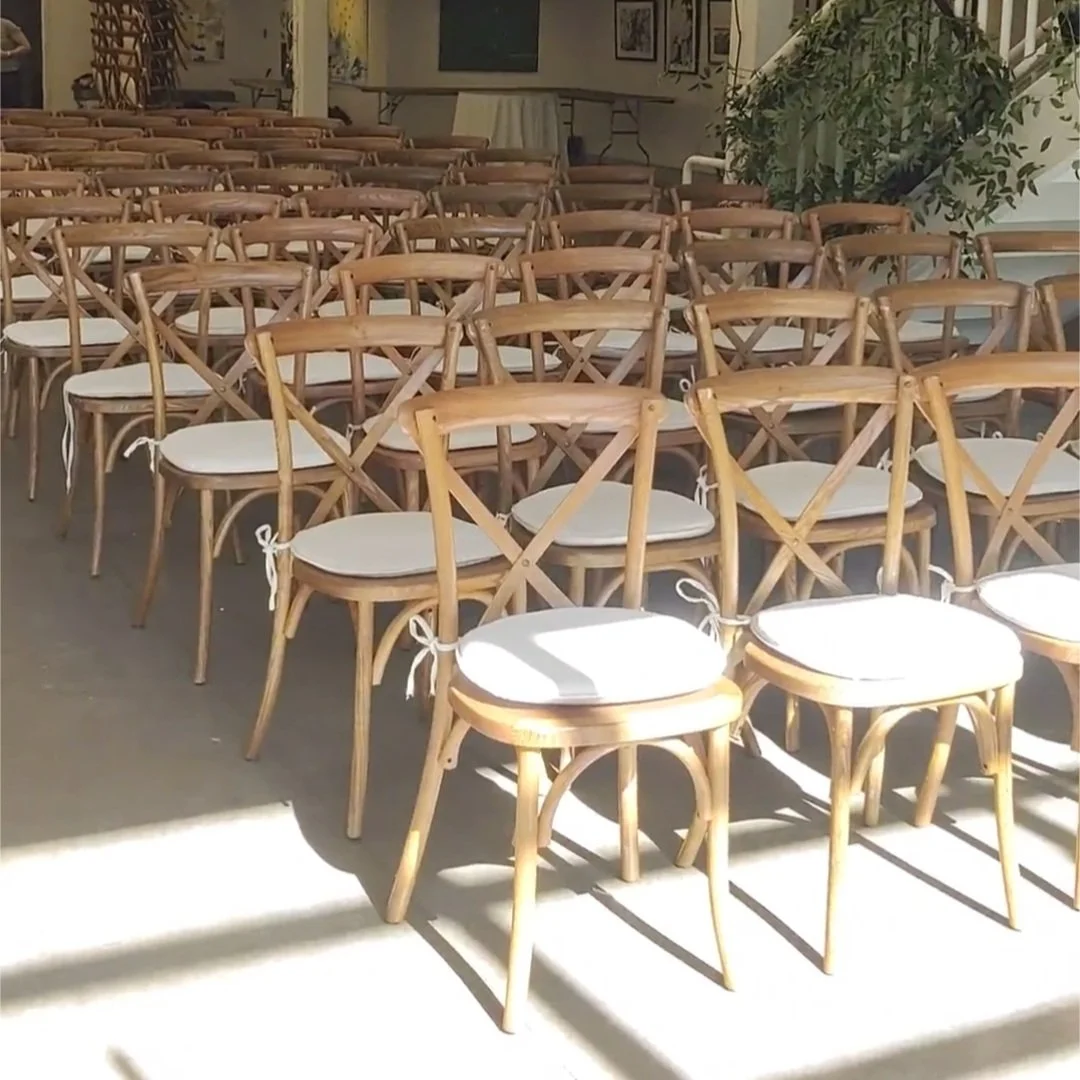 Multiple wooden chairs with white cushions arranged in rows in a well-lit room with artwork on the walls and a potted plant in the background.
