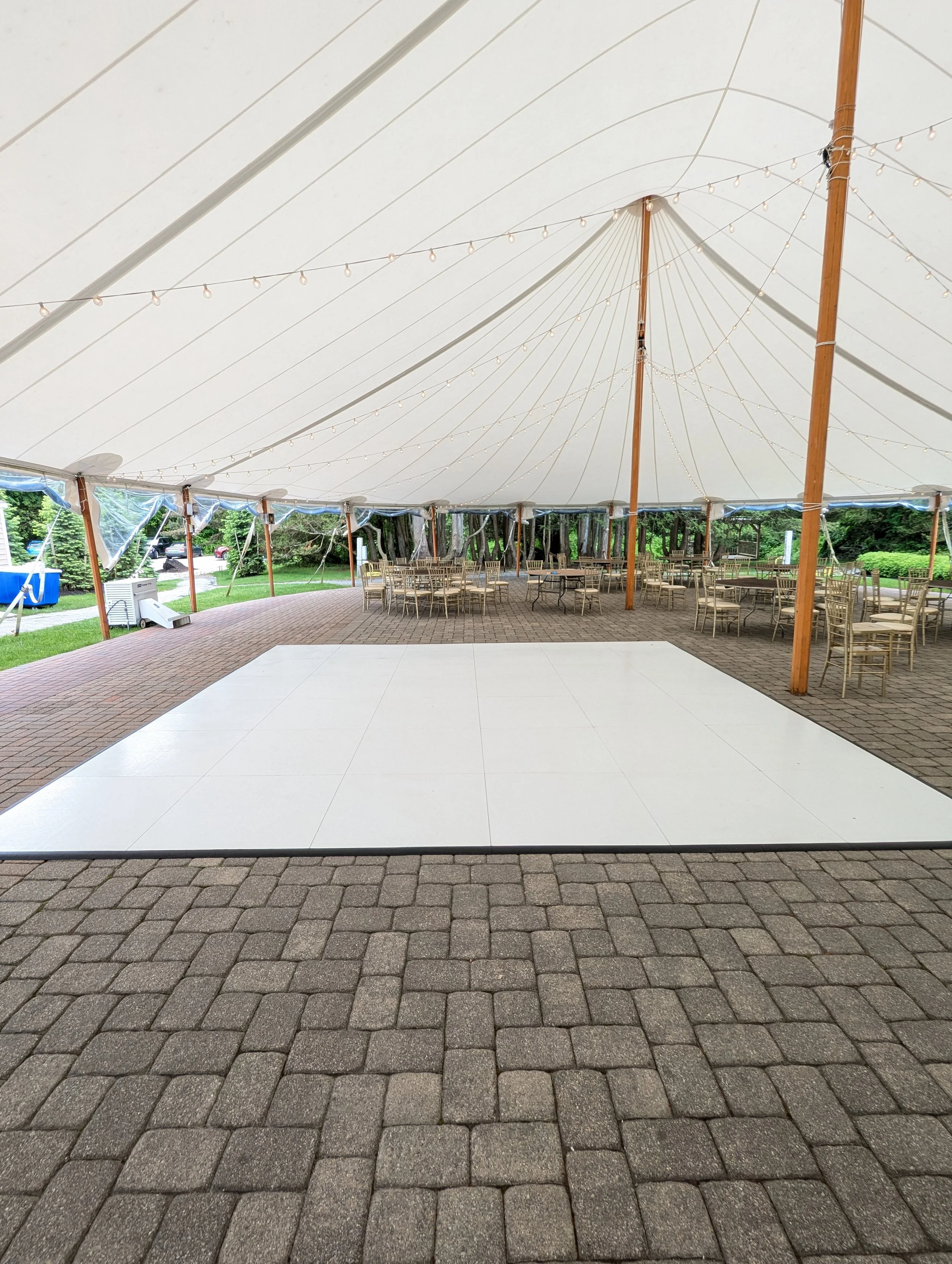 Empty outdoor event tent with string lights, gold chairs arranged around tables, and a white dance floor on brick paving, with greenery outside.