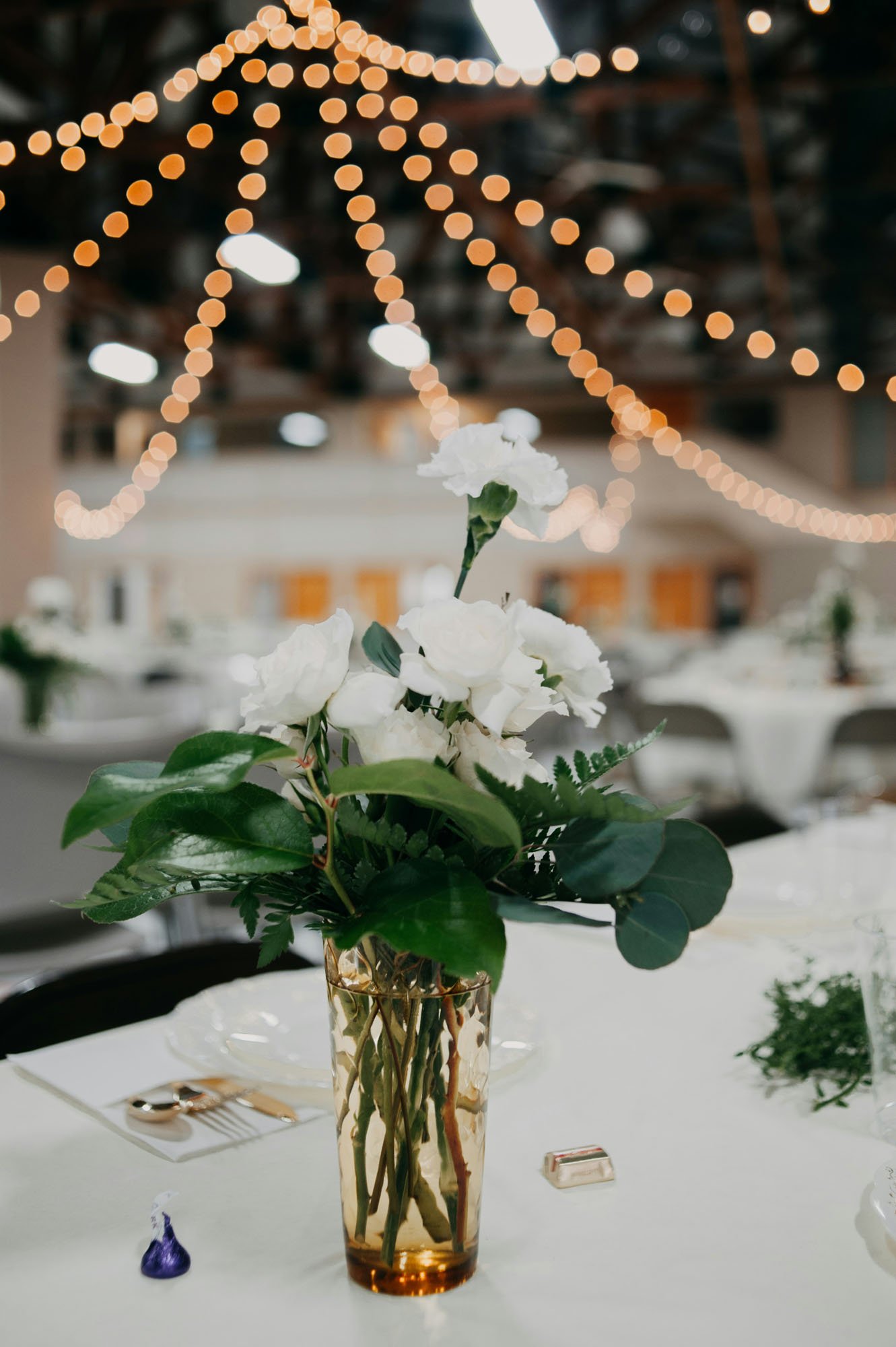 A floral centerpiece with white flowers and green leaves in a tall, amber glass vase on a table, decorated for an event with string lights overhead.