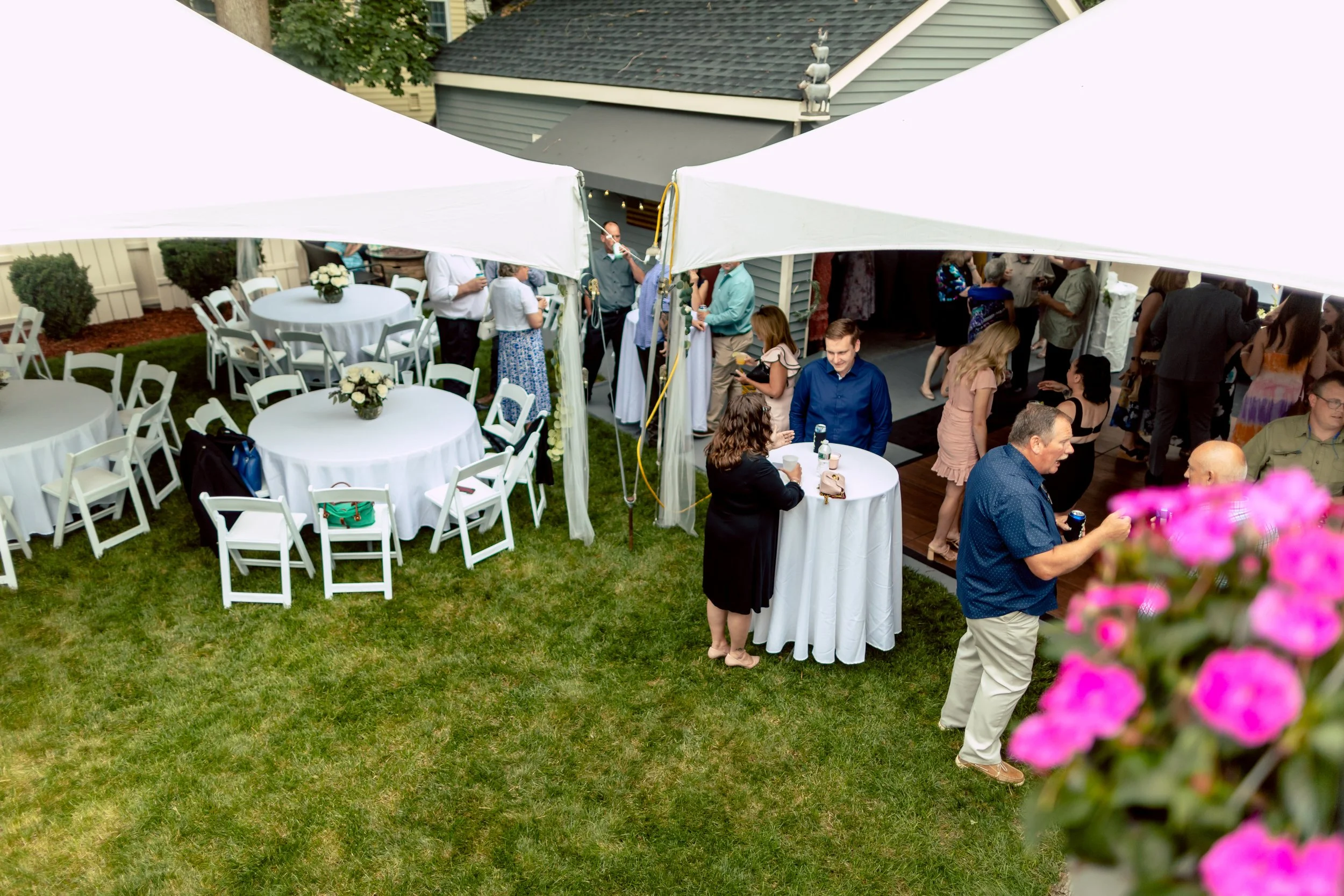 People socializing at a backyard outdoor event with round tables covered with white tablecloths, under a large white canopy. Some individuals are gathered near a tent, while others are dancing or conversing on the patio. The setting is a well-maintained yard with grass, flowers, and house siding visible in the background.