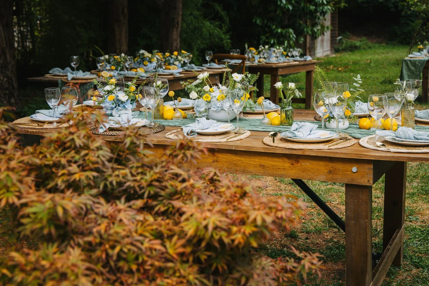 An outdoor dining table decorated with floral arrangements, lemon accents, and tableware, set in a lush garden during daytime.