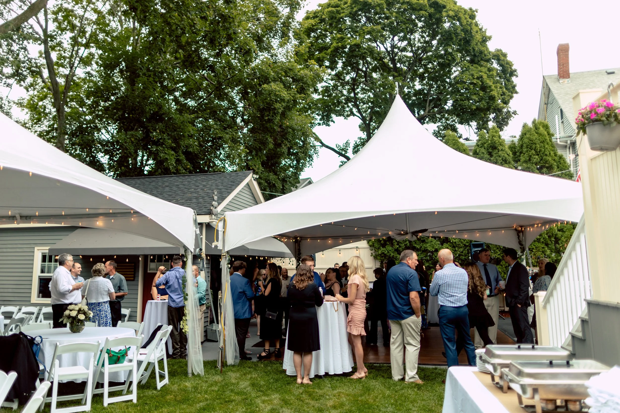 Guests socializing under white canopy tents at an outdoor party or event in a backyard with trees and houses in the background, tables with floral arrangements, and buffet stations.