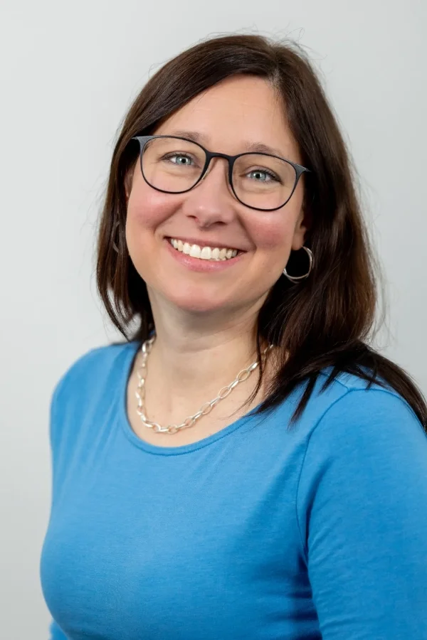 Lächelnde Frau mit brünettem Haar, Brillengläser, blauer Shirt und Schmuck, vor neutralem Hintergrund.