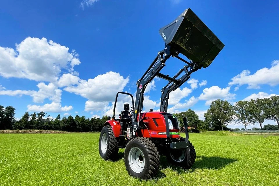 A red compact tractor with an upper lift attachment, parked on a green field under a blue sky with white clouds, and trees in the background.