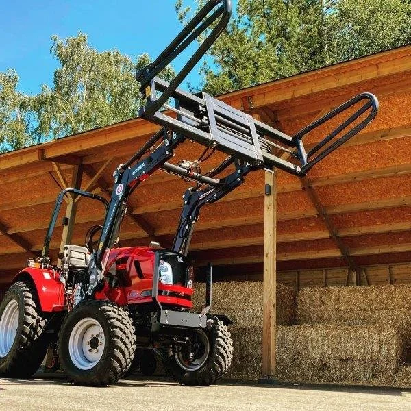 Knegt tractor, A red compact tractor with a front loader attachment lifting a metal platform with guardrails, located near hay bales under a wooden structure.