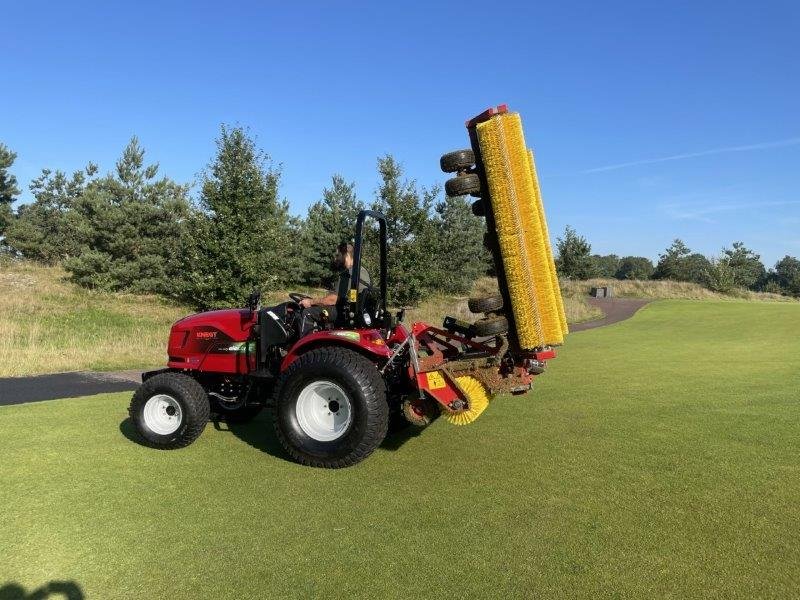 Knegt Tractor, A person on a red tractor with a yellow attachment for grooming a golf course, on a green field with trees and a clear blue sky.