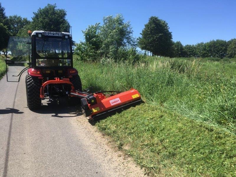 Knegt Tractor, A red and black tractor with a mower attachment cuts grass along the side of a rural road, with green trees and a clear blue sky in the background.