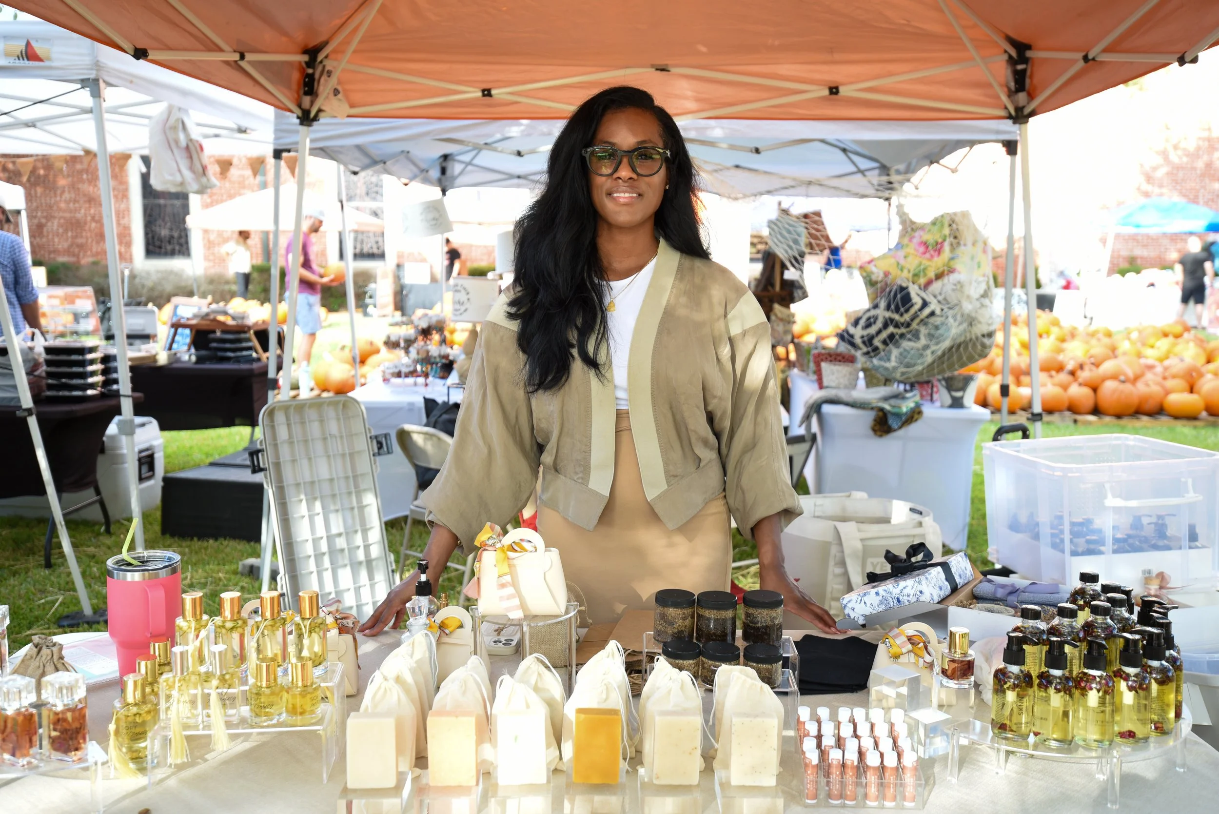 A woman with black hair and glasses smiling at a marketplace stall selling skincare products and perfumes. The stall is under orange and white tents, with other market stalls and pumpkins in the background.