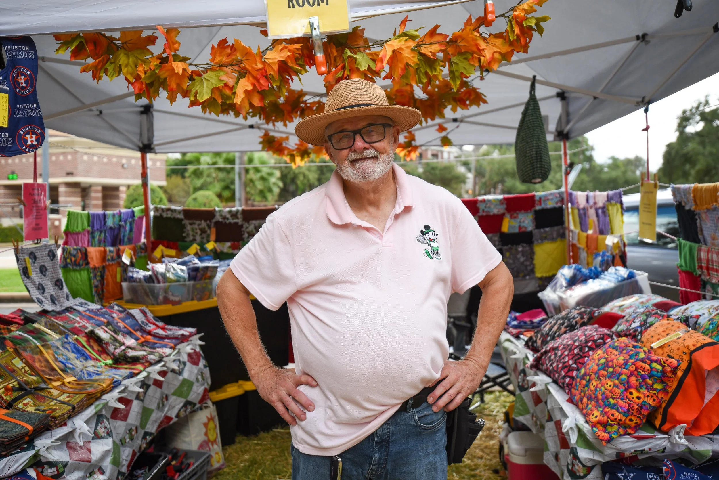 An older man with glasses and a hat standing at a craft fair booth under a white canopy, surrounded by colorful textiles and craft items.