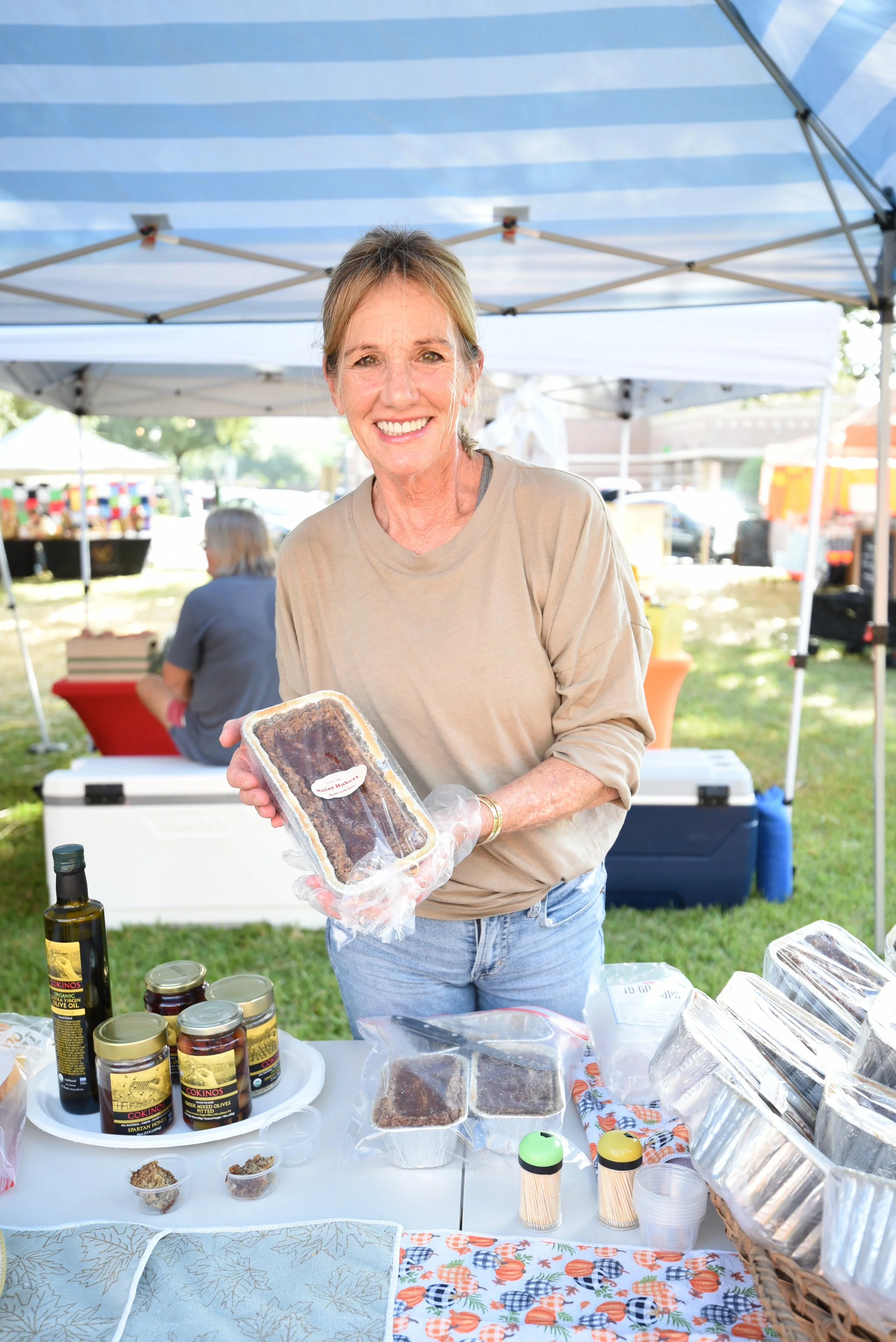 Smiling woman at outdoor market holding packaged food item, surrounded by jars, bottles, and food samples on table.