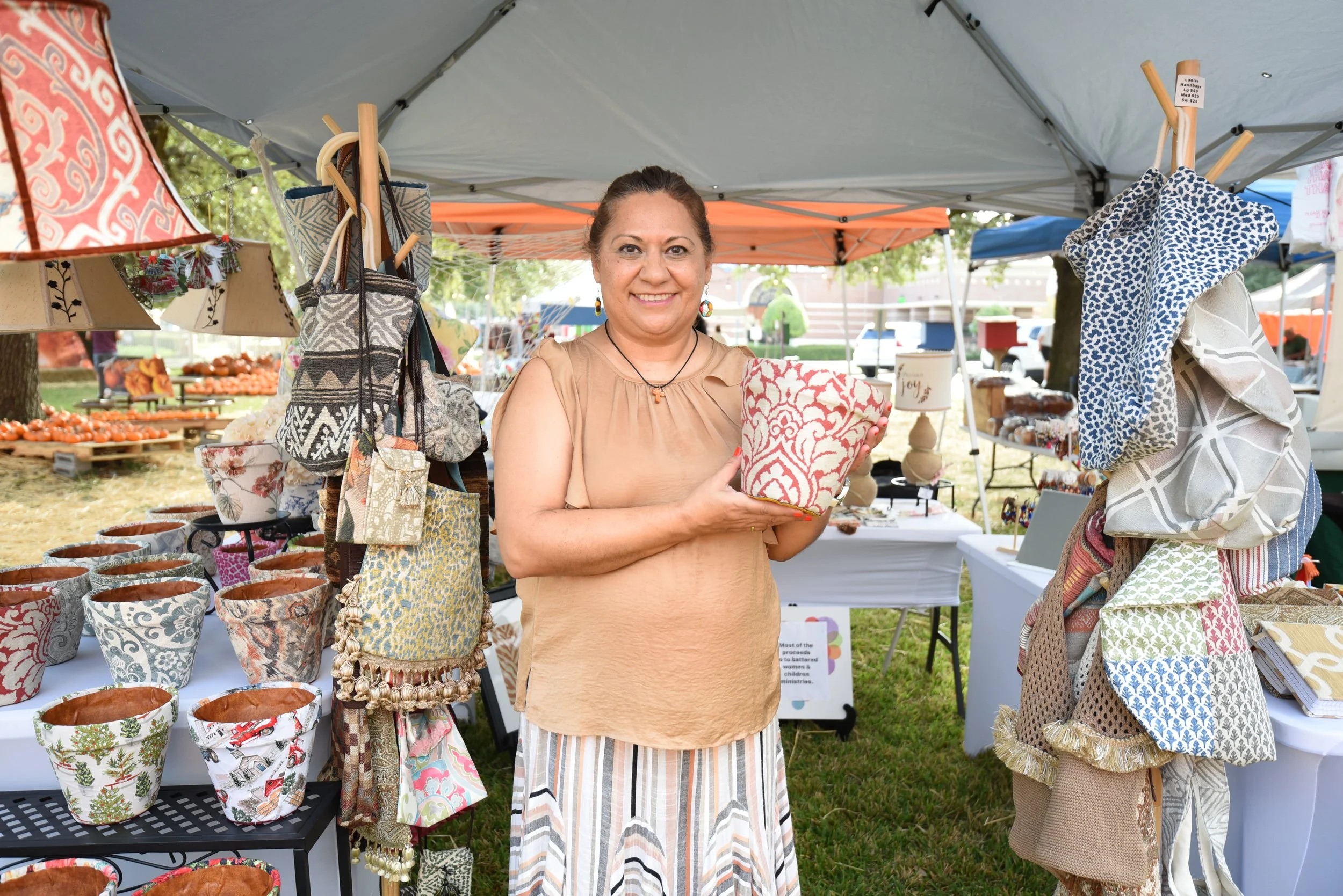 A woman standing at an outdoor market stall displaying woven bags and pottery.